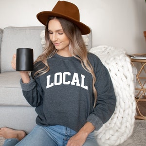 May include: A woman wearing a dark gray sweatshirt with the word "LOCAL" printed in white on the front. She is holding a black coffee mug and wearing a brown fedora.