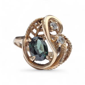 May include: A gold ring with an oval, dark teal gemstone at its centre. The ring has an ornate, openwork design with two smaller, clear gemstones. The ring's band is a simple, polished gold.