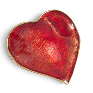 Puede incluir: Un plato en forma de corazón con un acabado de esmalte rojo vibrante y translúcido que muestra una capa base metálica texturizada. El objeto está delineado con un borde fino de metal pulido en tono dorado, creando un contorno suave y curvo.