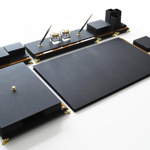 May include: Black leather desk organizer set with gold accents. The set includes a large writing surface, a smaller writing surface, a pen holder, a notepad holder, and a set of coasters.
