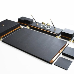 May include: Black leather desk organizer set with gold accents. The set includes a large writing pad, a pen holder, two inkwells, a letter tray, and a small tray.