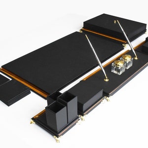 May include: Black leather desk organizer set with gold accents. The set includes a desk pad, pen holders, a letter tray, and inkwells.