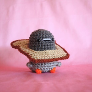 May include: A gray crocheted amigurumi toy wearing a brown and beige crocheted hat. The toy has black eyes and a white mouth.