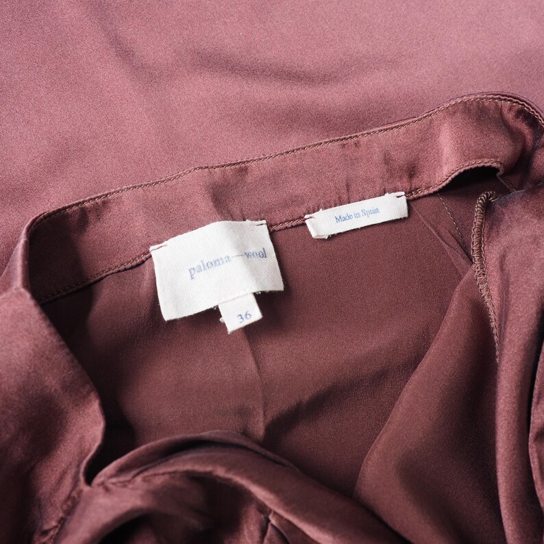 May include: A dark mauve garment with a smooth, satin-like finish. The interior features a white fabric label with the text 'paloma wool' in blue, a smaller tag below it with the number '36', and a separate tag reading 'Made in Spain'.
