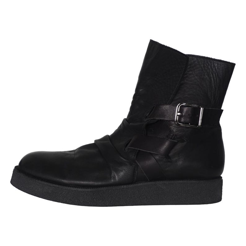 May include: A black ankle boot featuring a smooth leather upper, a thick textured rubber sole, and a decorative strap across the midfoot. The boot includes a side strap with a silver-toned metal buckle closure near the top of the shaft.