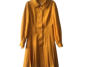 May include: A mustard yellow coat featuring a structured collar, long sleeves with buttoned cuffs, and a front button closure. The garment has a fitted waistline that transitions into a pleated skirt section, creating a flared silhouette.