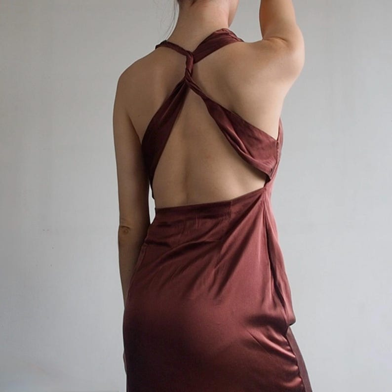 May include: A dark brown satin dress featuring a sleeveless design with a deep open back. The bodice includes a twisted strap detail that connects at the center of the back, creating a cutout effect above the waistline with a smooth, flowing skirt.