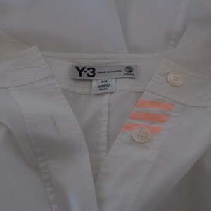 May include: White collared shirt with the Y-3, Yohji Yamamoto, and Adidas logos on the tag. The shirt has two buttons and three orange stripes on the right side. The shirt is labeled M/M and Made in China.