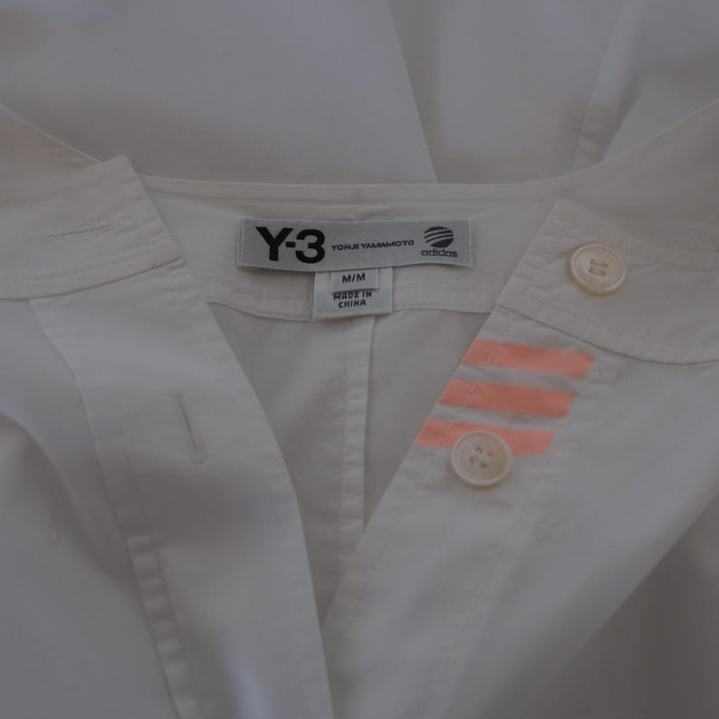 May include: White collared shirt with the Y-3, Yohji Yamamoto, and Adidas logos on the tag. The shirt has two buttons and three orange stripes on the right side. The shirt is labeled M/M and Made in China.