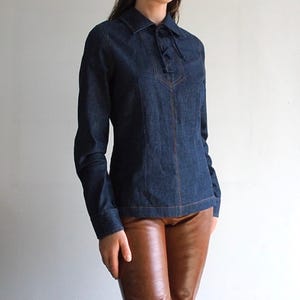May include: A dark blue denim shirt with long sleeves and a collared neckline. The shirt features a tie closure at the neckline and contrasting stitching. The shirt is paired with brown leather pants.