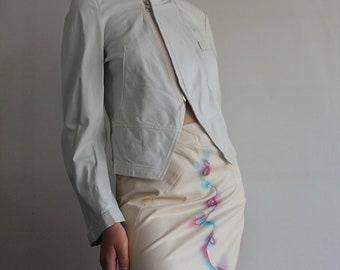 May include: A white leather jacket with a zipper closure and a collar. The jacket is cropped and has a slightly asymmetrical design. The model is wearing a white skirt with a colorful abstract design.