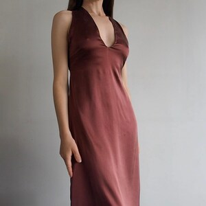 May include: A sleeveless midi-length dress in a deep mahogany brown color. The garment features a deep V-neckline, a fitted bodice, and a smooth, satin-like fabric with a subtle sheen that flows down into a straight, unadorned skirt silhouette.