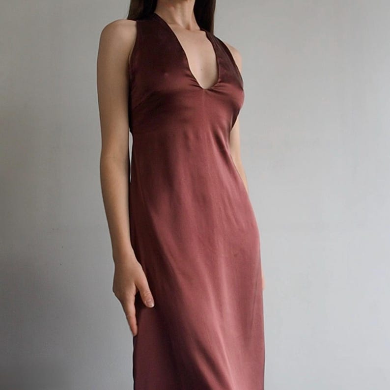 May include: A sleeveless midi-length dress in a deep mahogany brown color. The garment features a deep V-neckline, a fitted bodice, and a smooth, satin-like fabric with a subtle sheen that flows down into a straight, unadorned skirt silhouette.