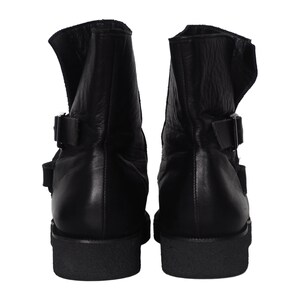 May include: A pair of black leather ankle boots featuring a high-top silhouette with two adjustable side straps and metal buckles. The boots have a thick, textured black rubber sole and a smooth leather upper with visible natural grain texture.