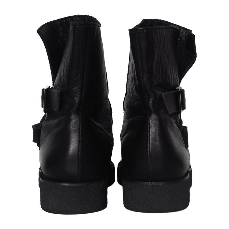 May include: A pair of black leather ankle boots featuring a high-top silhouette with two adjustable side straps and metal buckles. The boots have a thick, textured black rubber sole and a smooth leather upper with visible natural grain texture.