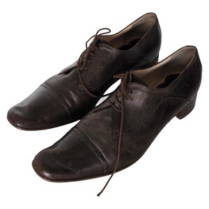 May include: A pair of dark brown leather dress shoes with a lace-up design. The shoes have a rounded toe and a low heel. The leather appears worn, giving them a vintage look. The laces are the same colour as the shoes.