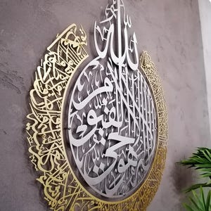 Op de afbeelding: Een groot, cirkelvormig wandkunstwerk met ingewikkelde Arabische kalligrafie. Het ontwerp bestaat uit een zilverkleurig middengedeelte omringd door een goudkleurige buitenrand, beide met gedetailleerde, lasergesneden schriftpatronen.