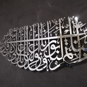 May include: A silver metal wall art piece featuring intricate Arabic calligraphy. The design is a traditional Islamic art form.