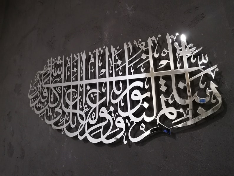 May include: A silver metal wall art piece featuring intricate Arabic calligraphy. The design is a traditional Islamic art form.