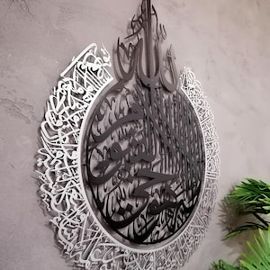 Op de afbeelding: Een cirkelvormig wandkunstobject met complexe Arabische kalligrafie. Het ontwerp bestaat uit een zwart middengedeelte met gedetailleerd schrift, omringd door een zilverkleurige buitenring van sierlijke, vloeiende Arabische letters.