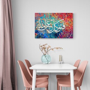 May include: A colorful abstract painting with Arabic calligraphy in white. The painting is framed and hanging on a white wall above a white table with two pink chairs.