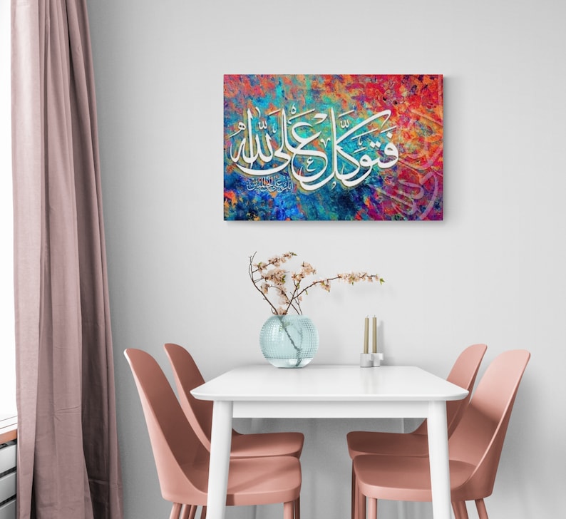 May include: A colorful abstract painting with Arabic calligraphy in white. The painting is framed and hanging on a white wall above a white table with two pink chairs.