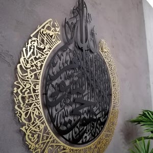 Op de afbeelding: Een cirkelvormig wandkunstobject met ingewikkelde Arabische kalligrafie. Het ontwerp bestaat uit een centrale zwarte metalen laag met gedetailleerd schrift, omringd door een grotere goudkleurige metalen rand met uitgebreide kalligrafische patronen.