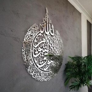 Op de afbeelding: Een groot, rond wandkunstobject van zilverkleurig metaal met ingewikkelde, lasergesneden Arabische kalligrafie. Het reflecterende oppervlak toont gedetailleerde schriftpatronen die een druppelvormig silhouet vormen op een grijze, getextureerde muur.