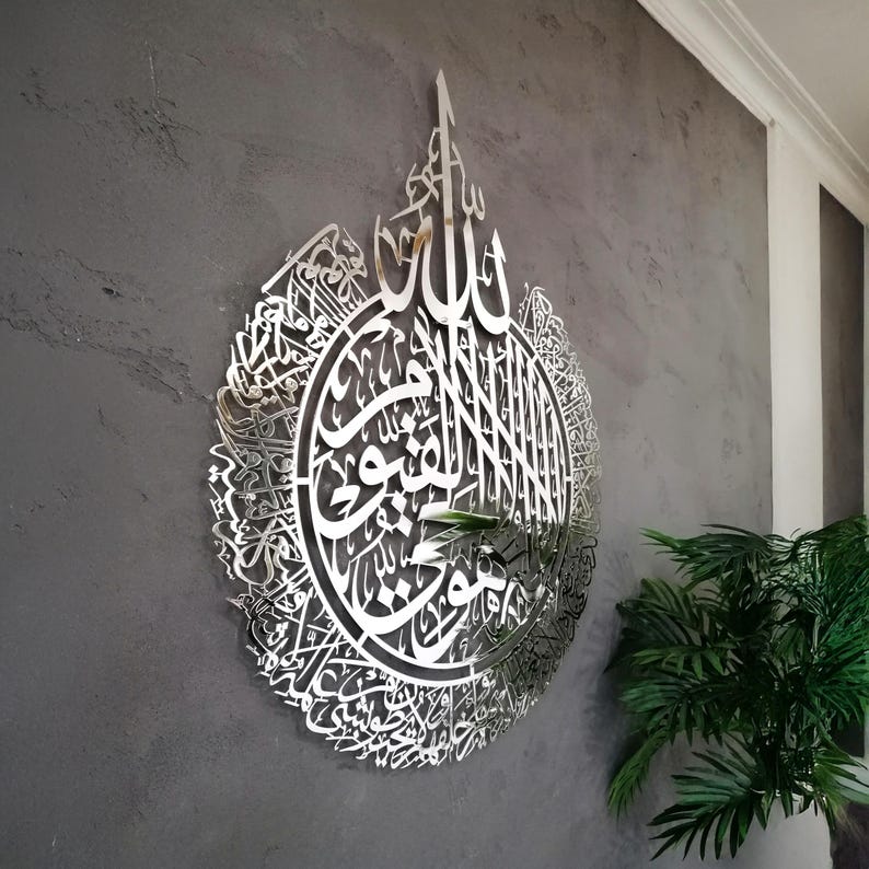 Op de afbeelding: Een groot, rond wandkunstobject van zilverkleurig metaal met ingewikkelde, lasergesneden Arabische kalligrafie. Het reflecterende oppervlak toont gedetailleerde schriftpatronen die een druppelvormig silhouet vormen op een grijze, getextureerde muur.