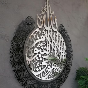 Op de afbeelding: Een cirkelvormig wandkunstwerk met complexe Arabische kalligrafie. Het midden is een gepolijste zilverkleurige metalen uitsnede met gedetailleerd schrift, omringd door een bredere zwarte metalen rand met dichte, sierlijke Arabische letters.