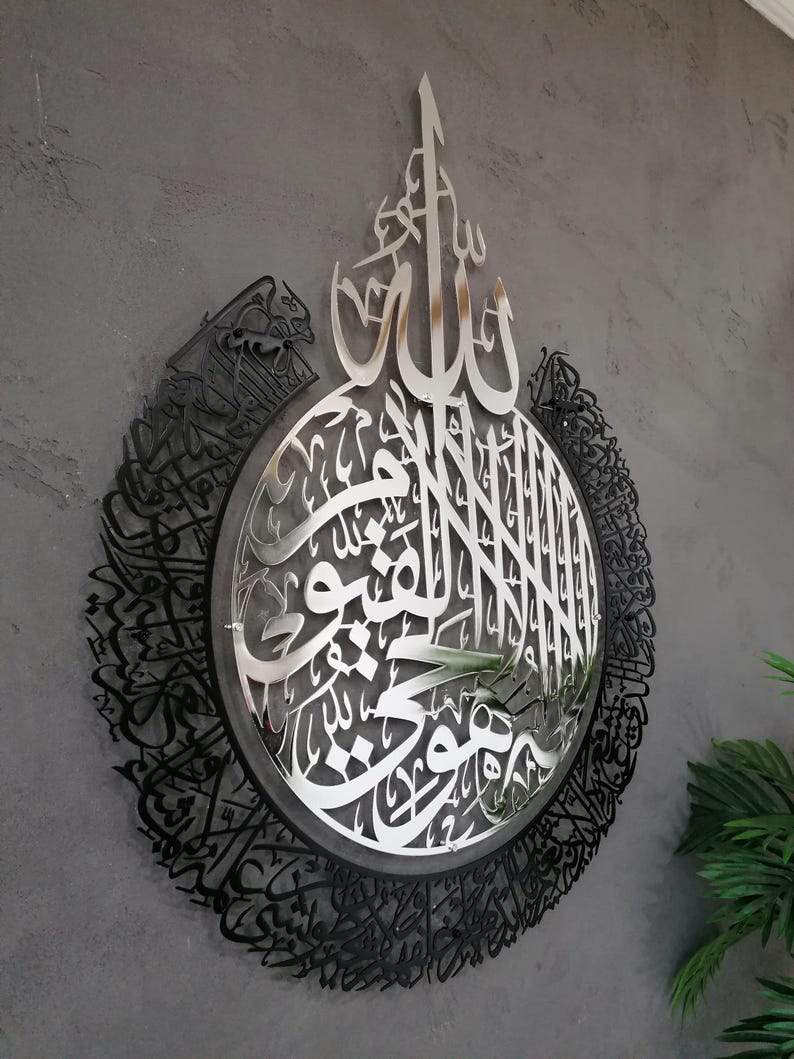 Op de afbeelding: Een cirkelvormig wandkunstwerk met complexe Arabische kalligrafie. Het midden is een gepolijste zilverkleurige metalen uitsnede met gedetailleerd schrift, omringd door een bredere zwarte metalen rand met dichte, sierlijke Arabische letters.