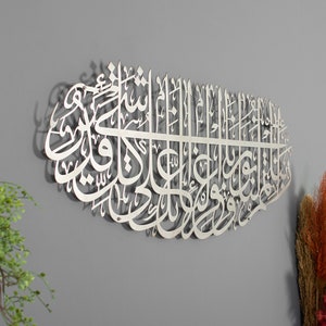 May include: A silver metal wall art piece featuring intricate Arabic calligraphy. The calligraphy is in a traditional style and is arranged in a circular pattern. The art piece is mounted on a gray wall.