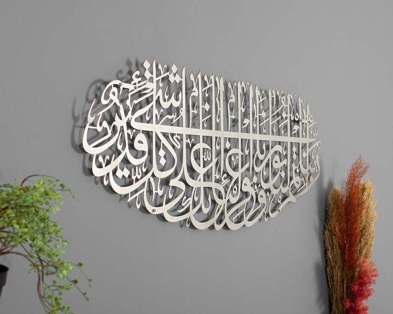 May include: A silver metal wall art piece featuring intricate Arabic calligraphy. The calligraphy is in a traditional style and is arranged in a circular pattern. The art piece is mounted on a gray wall.