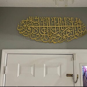 May include: A gold-colored metal wall decor featuring Arabic calligraphy in a decorative oval shape. The decor is mounted on a gray wall above a white door.
