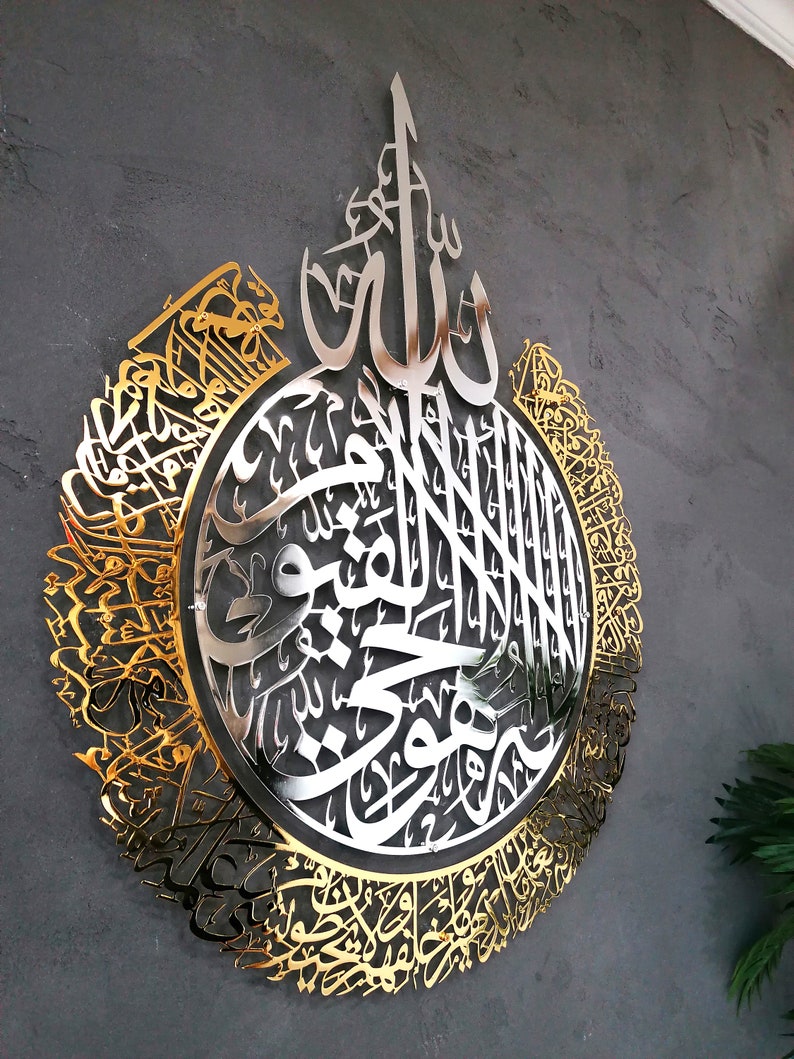 Op de afbeelding: Een groot, cirkelvormig wandkunstwerk met ingewikkelde Arabische kalligrafie in een gepolijste gouden afwerking. Het ontwerp bevat een zilverkleurig middengedeelte met gedetailleerd schrift, omringd door een sierlijke, filigraan gouden rand.