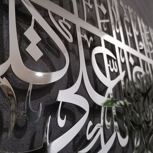 May include: A silver metal wall art piece with intricate Arabic calligraphy. The art is mounted on a dark gray textured surface.