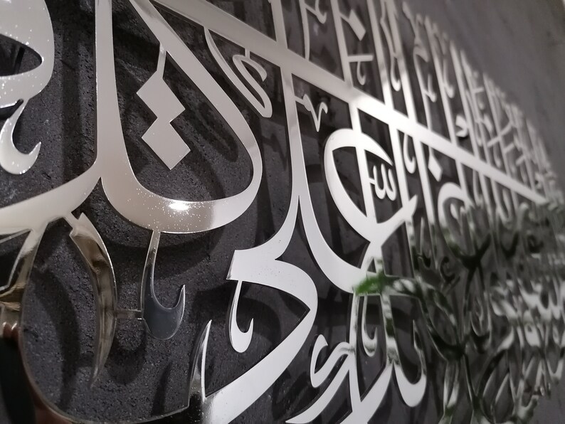 May include: A silver metal wall art piece with intricate Arabic calligraphy. The art is mounted on a dark gray textured surface.