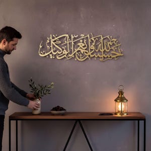 May include: A gold-colored metal wall art piece featuring intricate Arabic calligraphy script mounted on a gray wall. The design consists of flowing, interconnected letters and characters that form a horizontal, decorative display.