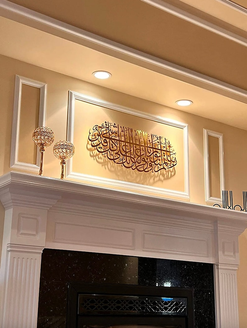 May include: A gold metal wall art piece with Arabic calligraphy, hanging above a white fireplace mantel with a black granite surround. Two gold candle holders with clear glass globes are on the mantel.