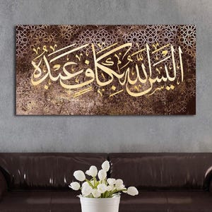 Op de afbeelding: Een rechthoekig canvas kunstwerk met gouden Arabische kalligrafie op een getextureerde bruine achtergrond met een decoratief patroon. Het kunstwerk wordt weergegeven boven een bruine leren bank, met een wit bloemstuk ervoor.