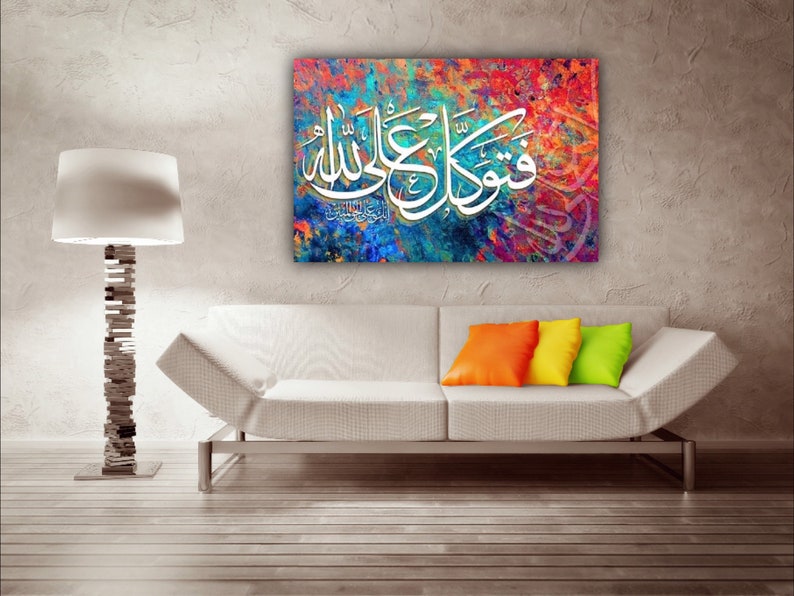 May include: A colorful abstract painting with white Arabic calligraphy. The painting is framed and hanging on a wall above a white couch with orange and yellow throw pillows.