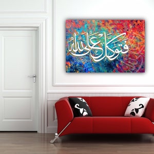 May include: A colorful abstract canvas print with white Arabic calligraphy that reads "Subhana Allahi wa bihamdihi, Subhana Allahi al-Azim". The print is hanging on a white wall above a red couch.
