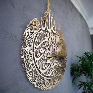 Op de afbeelding: Een groot, cirkelvormig wandkunstobject van goudkleurig metaal met ingewikkelde, lasergesneden Arabische kalligrafie. Het ontwerp is zeer gedetailleerd met sierlijke, vloeiende schriftpatronen die een samenhangend en decoratief geheel vormen.