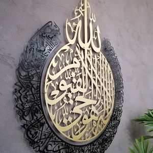 Op de afbeelding: Een cirkelvormig wandkunstobject met ingewikkelde Arabische kalligrafie. Het centrale deel is een goudkleurige metalen uitsnede, terwijl de buitenrand een zwart metalen frame is met gedetailleerd schrift. Het ontwerp is gelaagd en diep.