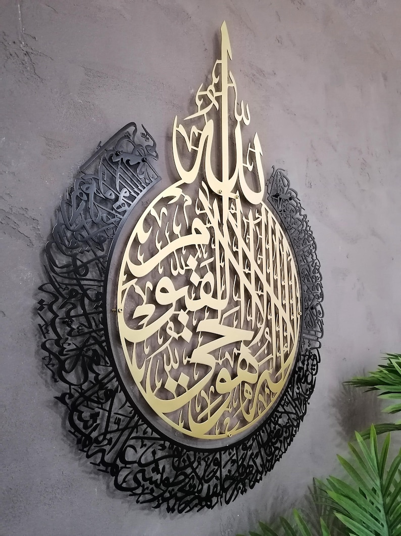 Op de afbeelding: Een cirkelvormig wandkunstobject met ingewikkelde Arabische kalligrafie. Het centrale deel is een goudkleurige metalen uitsnede, terwijl de buitenrand een zwart metalen frame is met gedetailleerd schrift. Het ontwerp is gelaagd en diep.