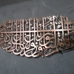 May include: A copper-colored metal wall art piece featuring intricate Arabic calligraphy. The design is a traditional Islamic art form.