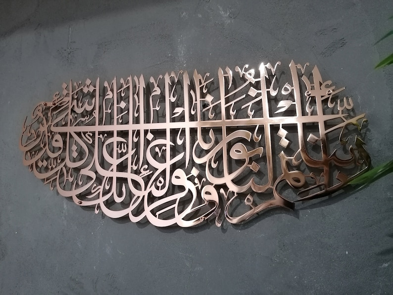 May include: A copper-colored metal wall art piece featuring intricate Arabic calligraphy. The design is a traditional Islamic art form.