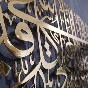 May include: Gold-colored metal wall art featuring intricate Arabic calligraphy. The design is a large, flowing script with a shiny, reflective finish.