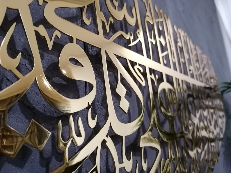 May include: Gold-colored metal wall art featuring intricate Arabic calligraphy. The design is a large, flowing script with a shiny, reflective finish.