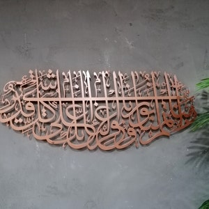 May include: A copper-colored metal wall art piece featuring intricate Arabic calligraphy. The design is in the shape of a crescent moon and features a repeating pattern of stylized letters.